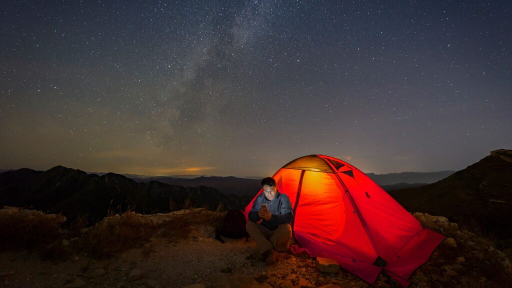 A camper using a phone beside a glowing red tent beneath a starry sky.
